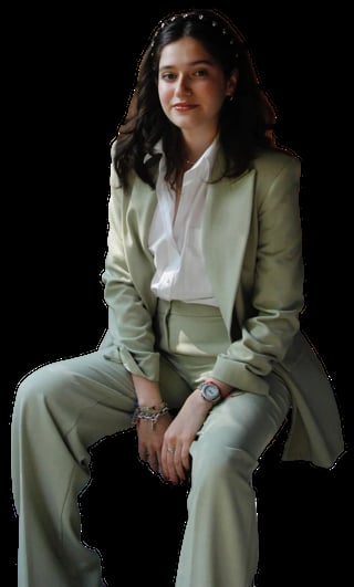Woman in sage green suit with white blouse, wearing jewelry and sitting against black background
